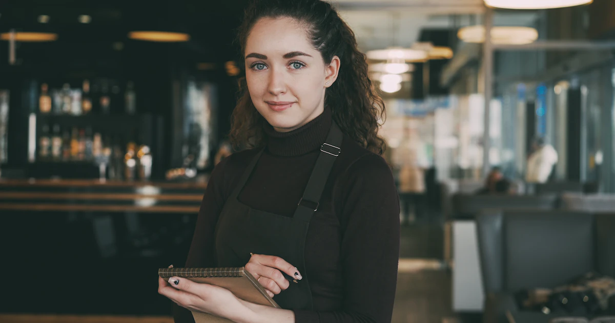 A restaurant server in an apron holding an order notepad in a busy dining room